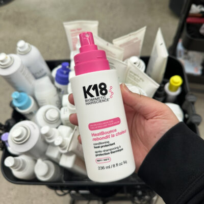 A sleek bottle of K18 HeatBounce Conditioning Heat Protectant being used by a professional stylist in a modern salon setting.
