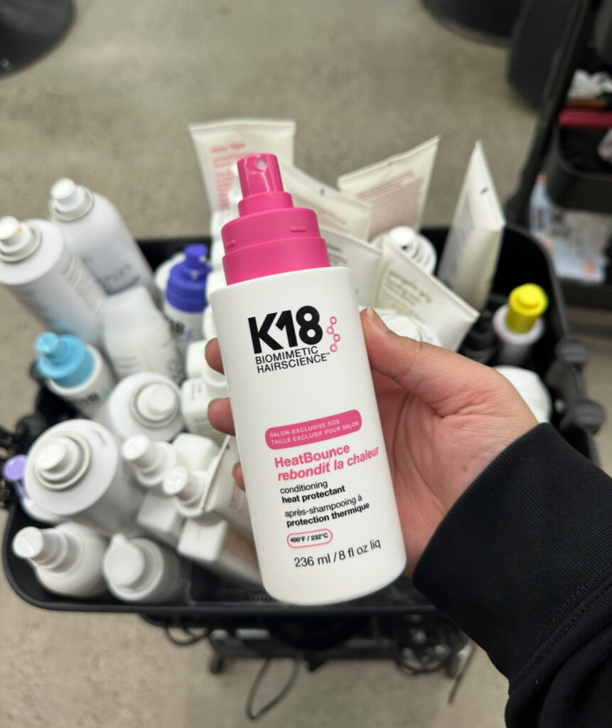 A sleek bottle of K18 HeatBounce Conditioning Heat Protectant being used by a professional stylist in a modern salon setting.